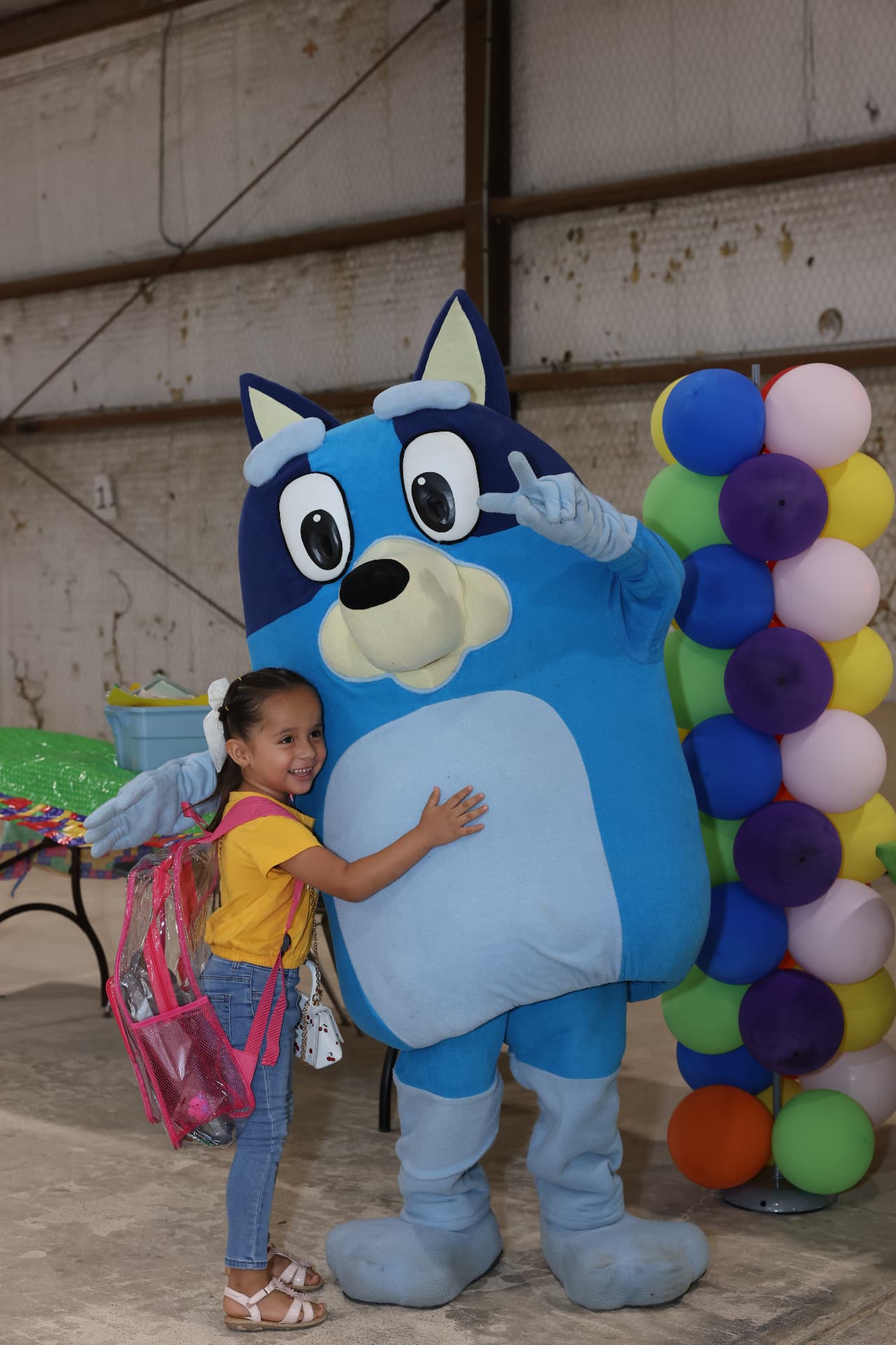 A young girl with a clear backpack hugging the Bluey mascot beside a balloon column.