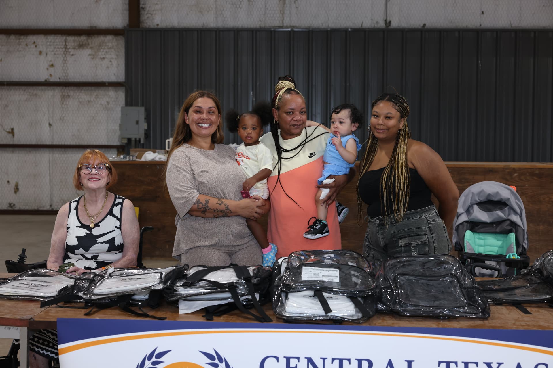 Central Texas Children's Academy volunteers with mothers and toddlers behind a table of clear backpacks.