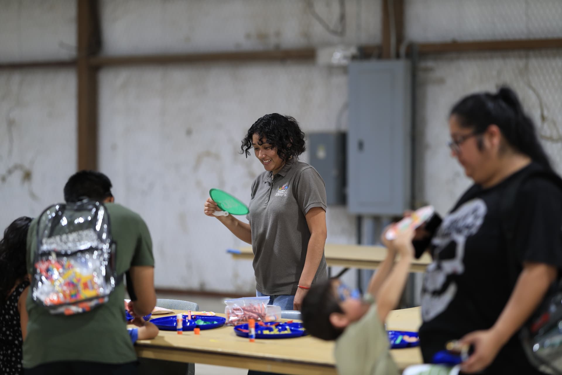 A volunteer handing out frisbees at the kids craft table as a child wears a clear backpack.