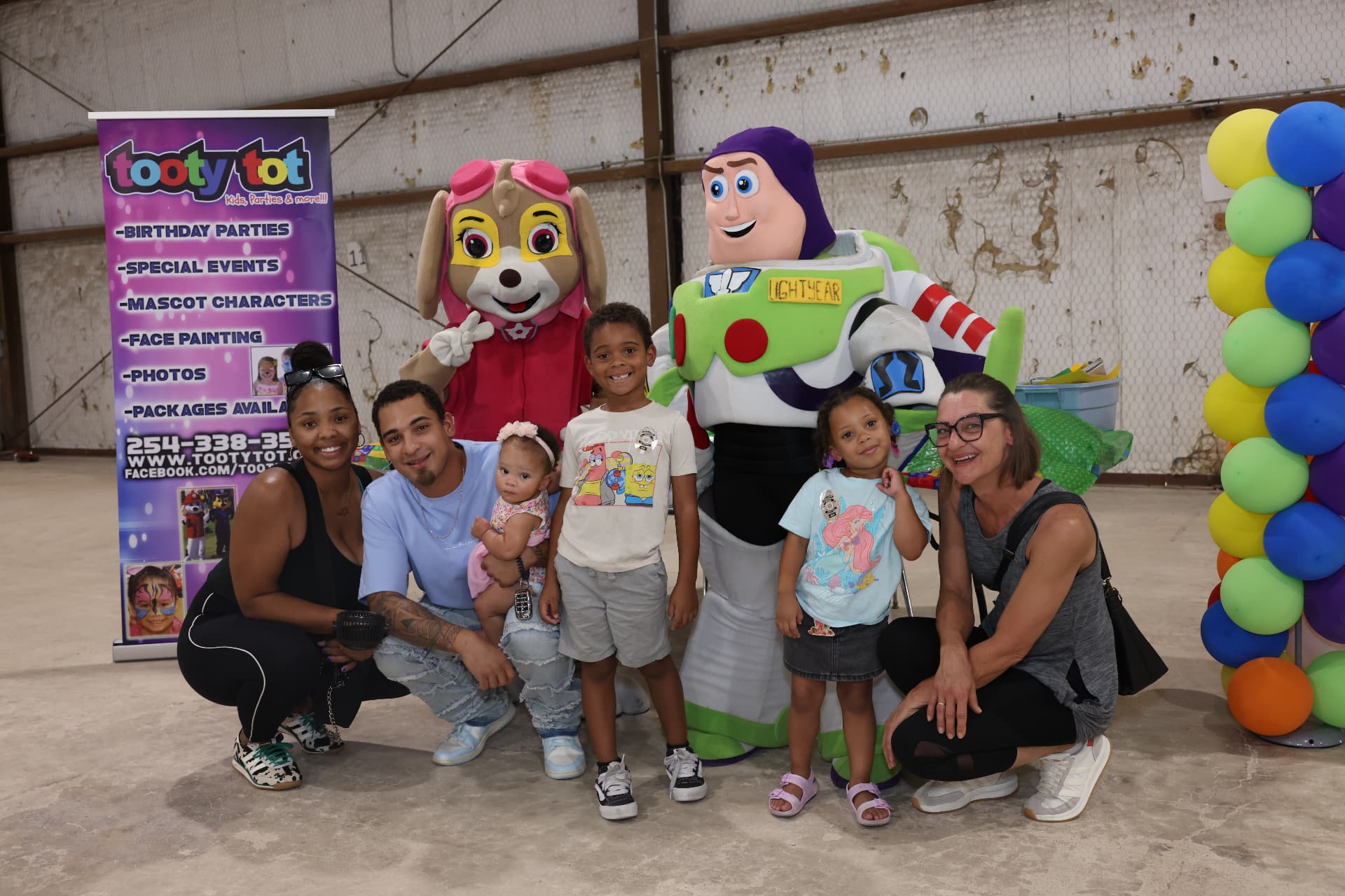 A family posing with Tooty Tot mascot characters at the photo backdrop.